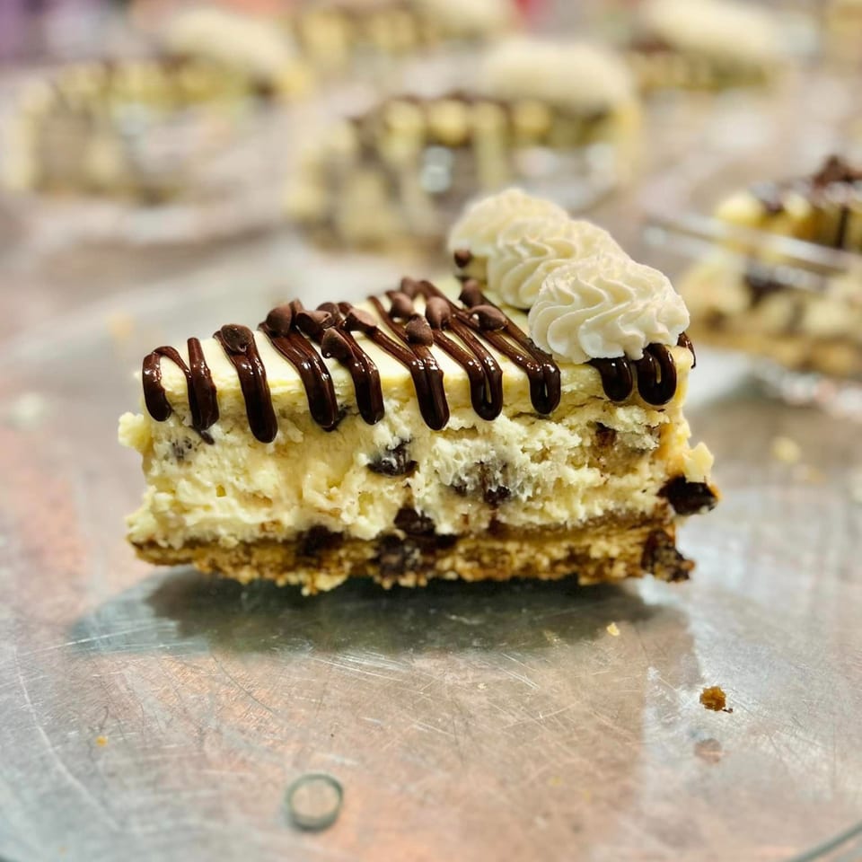 A slice of cheesecake with chocolate drizzle, chocolate chips, and whipped cream on a metal surface.